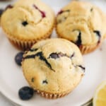 3 lemon blueberry muffins on a small white plate with fresh blueberries and lemon.