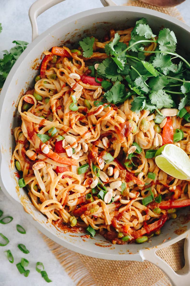 Peanut noodles in a pan with lime wedges, edamame and crushed peanuts.