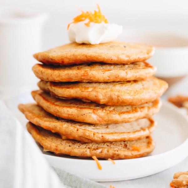 6 carrot cake pancakes stacked on top of each other in a white plate with yogurt and grated carrot.